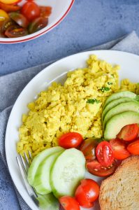 vegan egg scramble served in a white plate with cherry tomatoes, bread slice, and cucumber.