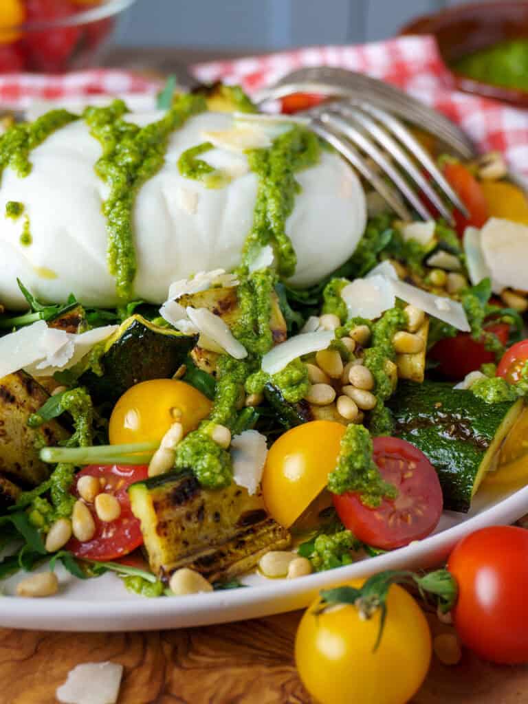 Close-up of burrata drizzled with pesto on a bed of arugula, cherry tomatoes, grilled zucchini, pine nuts, and shaved Parmesan.