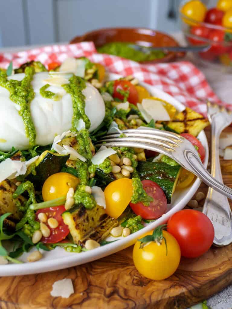 Overhead view of arugula burrata salad with pesto, cherry tomatoes, grilled zucchini, and pine nuts on a textured background with a red gingham napkin.