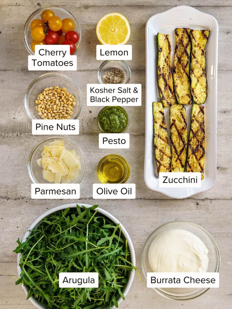 Labeled ingredients for arugula burrata salad arranged on a light surface, including cherry tomatoes, lemon, zucchini, arugula, burrata, pesto, pine nuts, Parmesan, olive oil, and seasoning.