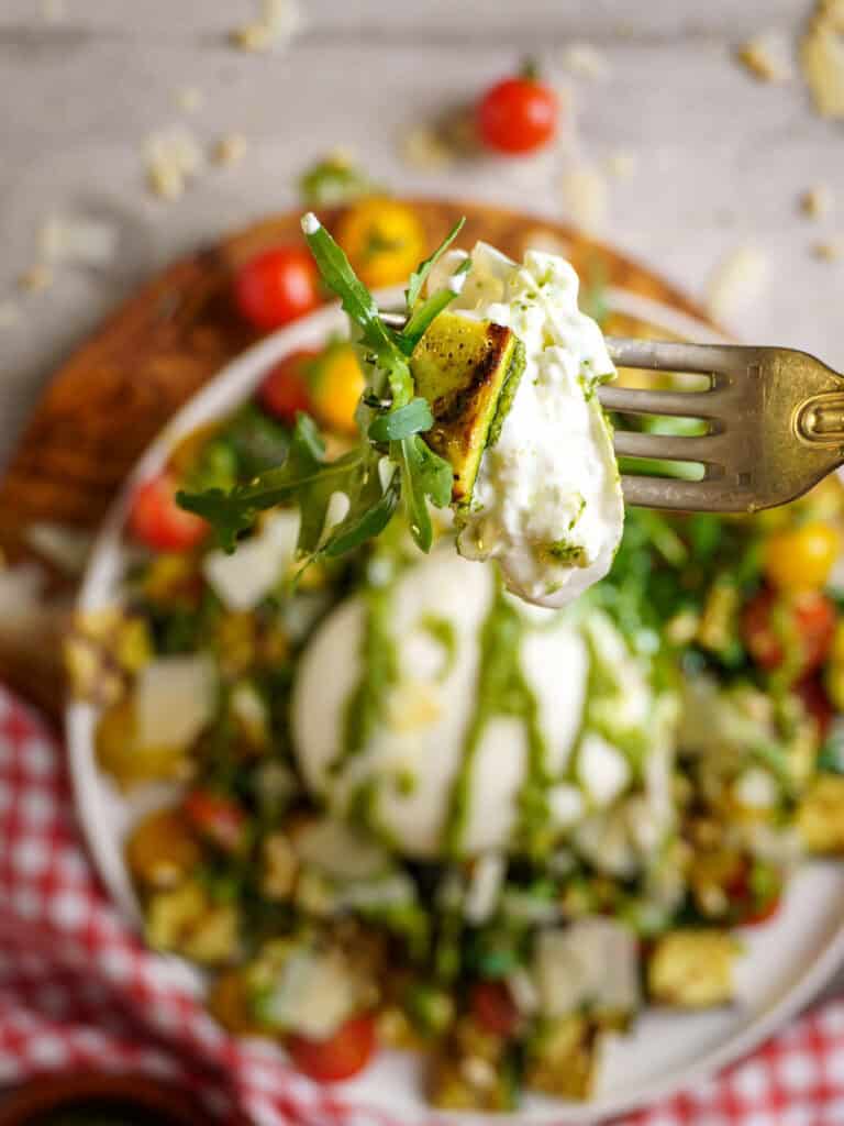 Forkful of creamy burrata, arugula, and grilled zucchini held above the plated salad with cherry tomatoes and scattered Parmesan in the background.