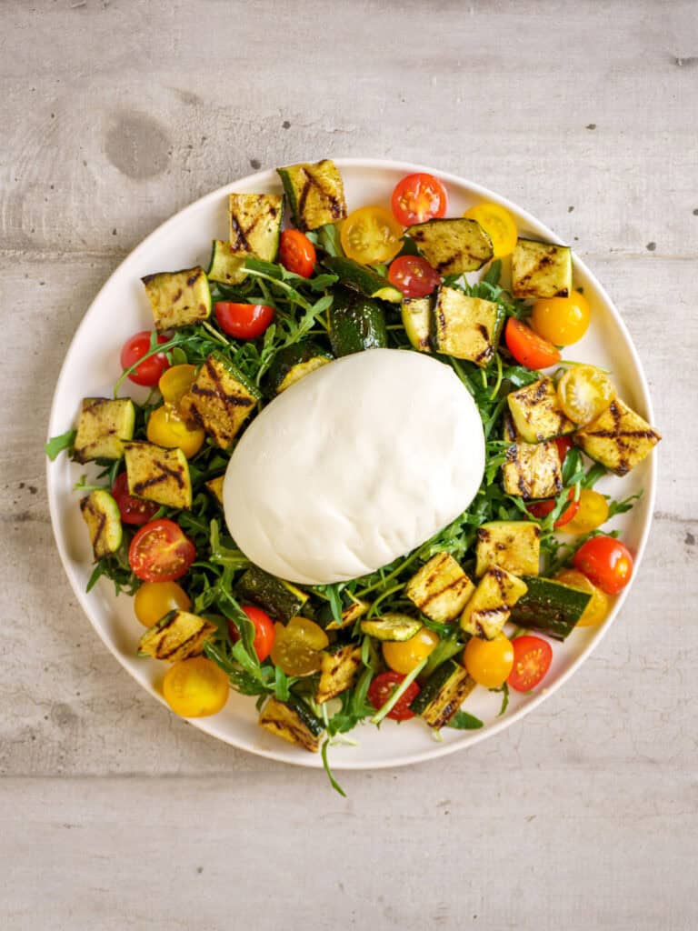 Burrata-topped salad fully assembled with pesto drizzled over and shaved Parmesan scattered across the plate.
