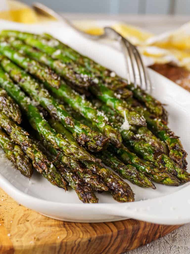 Charred asparagus laid on a white plate over a rustic wood board, with salt flakes and a yellow cloth in the background.