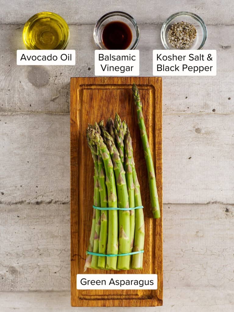 Overhead view of labeled ingredients for grilled asparagus: a bundle of green asparagus on a wooden board, with avocado oil, balsamic vinegar, kosher salt, and black pepper in glass bowls above.