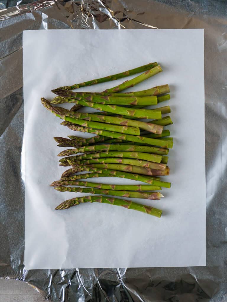 Raw asparagus spears arranged on a sheet of parchment paper placed over aluminum foil.