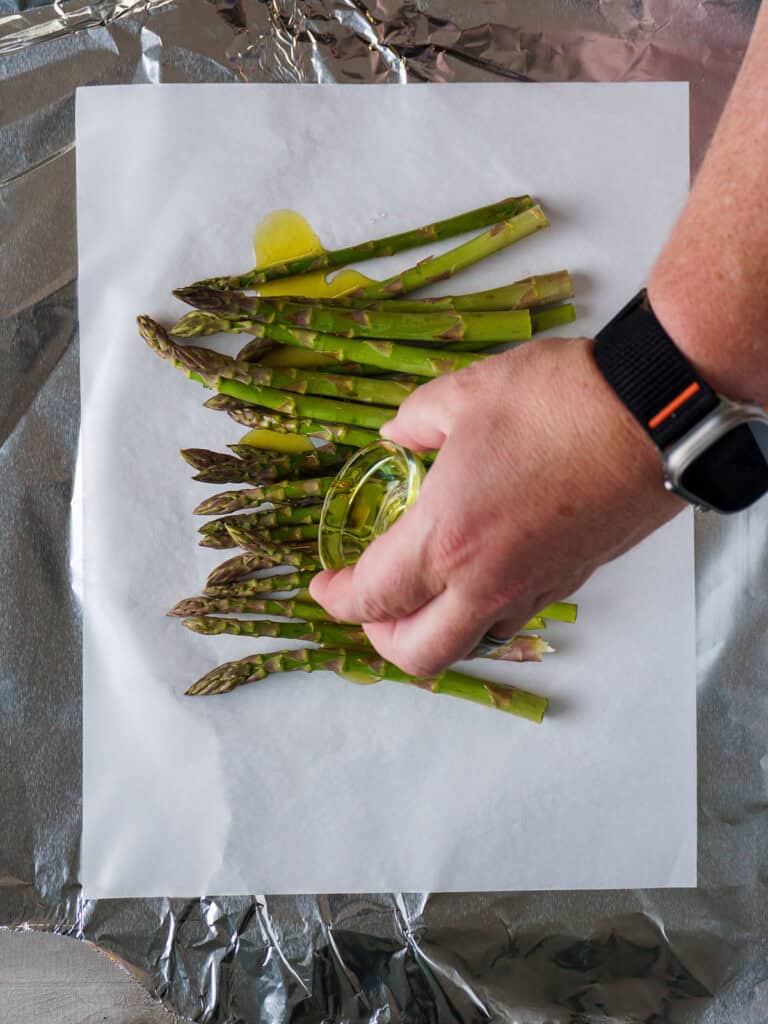 Hand pouring avocado oil over asparagus spears arranged on parchment and foil.