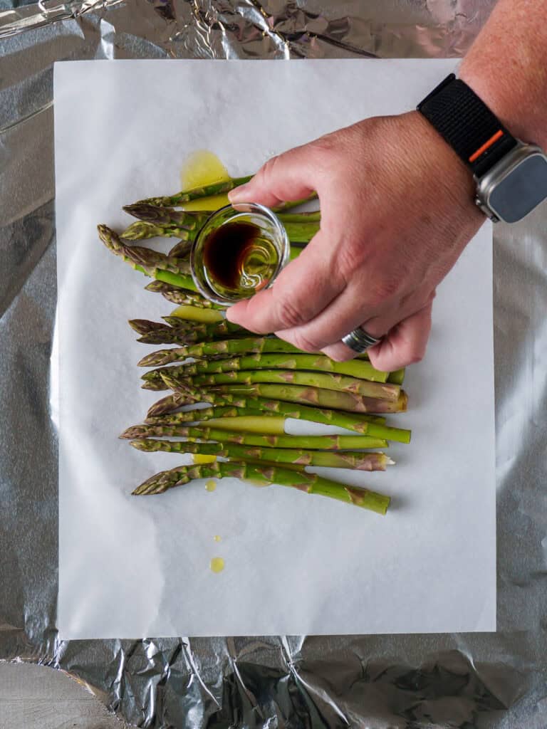 Hand holding a glass bowl of balsamic vinegar above the asparagus, ready to drizzle.