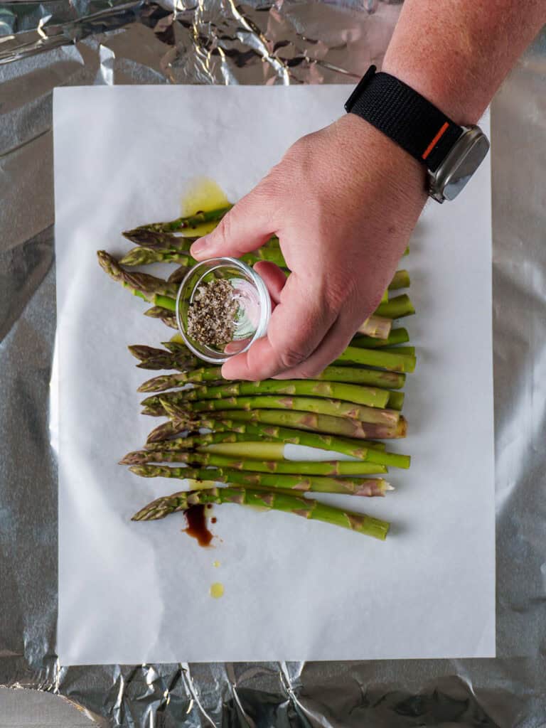 Hand seasoning asparagus with a small bowl of kosher salt and black pepper.
